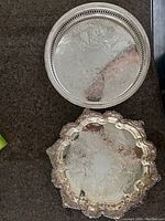 Two silver trays shown from top, detailed floral engravings, one with pierced rim, other with scalloped edge