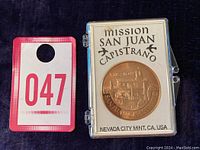 Front view of the Mission San Juan Capistrano collectible coin in clear plastic case with red numbered tag next to it for scale.