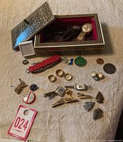 Overview of box with patterned lid open and contents displayed on surface including rings, cufflinks, pocket knife, pins, coins, and stones.
