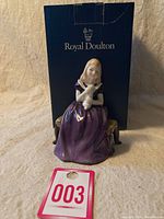 Figurine seated on small wooden stool holding white rabbit in purple dress with original Royal Doulton box behind it and tag 003 in front.