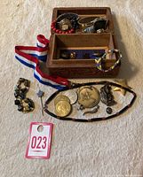 Photo showing assorted watches inside a carved wooden box, various coins, pins, medals, a star-shaped keychain pendant, a multicolor braided bracelet with button, and a tiger eye stone bead bracelet.