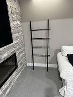 Black ash wood and steel blanket ladder standing empty beside white couch and stone fireplace on carpet.
