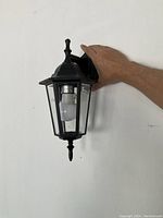 Front view of black carriage-style exterior light fixture with a person’s hand for size reference, showing glass panels, light bulb, and black metal frame.