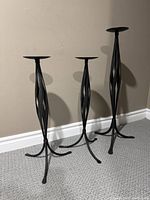 Front view of three black twisted iron floor standing candle holders of varying heights against beige wall and carpet floor.