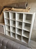 White IKEA KALLAX shelving unit with 16 square compartments, stacked with rolled rugs on top, against a concrete wall background, showing the full front view with all compartments visible.