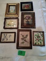 Seven small framed vintage artworks arranged on a white textile surface showing variety of floral and animal themes.