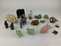 Full set of 7 vintage colored glass, ceramic and stone miniature animals displayed on white surface, showing various animals including elephant, rabbit, duck, dolphin, flamingo, lamb and lizard.