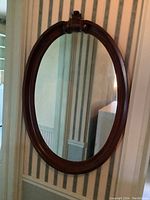 Full view of large oval mirror hanging against striped wallpaper background, showing overall shape and wood frame.