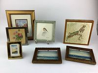 Seven miniature framed art pieces on white surface showing various subjects including a praying mantis, bird, flowers, seascapes, and architecture.
