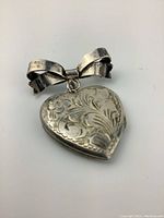 Close up photo of the vintage sterling silver heart shaped locket pin with engraved floral and swirl patterns attached to a bow-shaped pin.