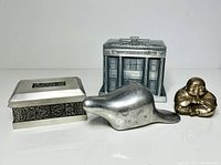 Four metal decor pieces in a group photo including pewter beaver, Royal Selangor box, metal commemorative money bank building, small Buddha figurine.