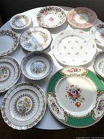 Top view of 11 assorted vintage porcelain plates with floral and scenic designs on a white table
