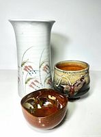 Three Asian-inspired pottery pieces with tall floral vase, footed cup with abstract glaze, and small brown bowl