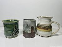 Three pottery pieces: green mug with tree, textured brown mug with painted trees, and white-brown jug