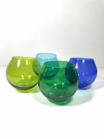 Four coloured Roly Poly glasses in lime green, light blue, dark green, and cobalt blue, placed on a white surface.