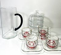 Clear hexagonal glass pitcher with black handle, four red-trimmed glass cups, matching square saucers, and hexagonal lidded glass container with compartments