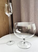 Two Moser giant snifter glasses of different shapes on a white table in front of a curtain and wood panel wall.