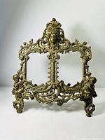 Full view of ornate brass double picture frame showing intricate scroll and floral design with central face embellishment.