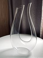 Full view of the Riedel Amadeo wine decanter showing the elegant double spout curved design, standing on a white surface with a backdrop of dark curtains.