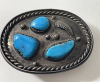 Front view of an oval sterling silver belt buckle decorated with three polished turquoise stones, small silver bead decorations, and a feather motif around the stones. The outer edge has a twisted rope design.