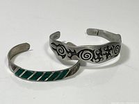 Two sterling silver bracelets side by side showing designs, one with green enamel stripes and the other with tribal etched design.