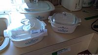 Four white CorningWare Cornflower Blue casserole dishes with glass lids except one missing lid, showing blue flower pattern, on kitchen counter