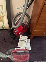 Red Dirt Devil vacuum on carpet with hose, metal wand, power cord visible alongside cleaning accessories still in plastic and manuals.