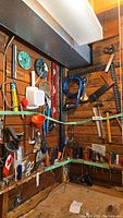 Tools hanging on wooden workshop wall including levels, squares, saws, hammers, scissors, and pliers arranged on hooks.