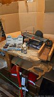 Full lot shot on wooden table with brown cardboard background showing miter box, miter table, sanding drum set, and hand tools.
