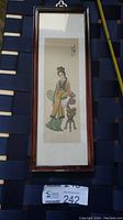 Full view of framed Japanese watercolor painting depicting woman in traditional clothing holding fan, standing next to ornamental stand.