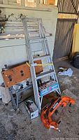 Overall view showing the aluminum ladder leaning against a cabinet, the orange Workmate collapsible work bench and steel sawhorse frames on the floor.