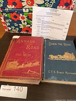 Two hardcover antique books side by side on a table with a floral background, showing front covers and a printed sheet with information about the books.