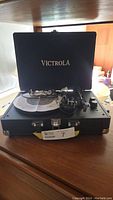 Open black suitcase-style Victrola turntable showing turntable platter, tonearm, control knobs and cable coiled inside, with lid open showing Victrola logo.