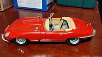 Side view of the red 1961 E Type Jaguar model car on a wooden surface showing intact frame but worn condition.