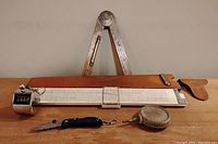 Vintage slide rule with moving cursor and attached mechanical counter, wooden expandable carpenter's ruler in background, small black tool and round tape measure at front