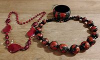Photo showing two costume jewelry necklaces and one bangle bracelet placed on a wooden surface. The necklaces are red based with one featuring large translucent red beads and the other red and black patterned beads resembling snakeskin. The bangle is black with red designs.