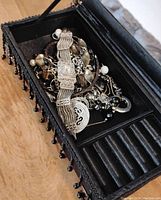 Jewelry box open showing assorted silver-tone costume jewelry including a wristwatch with metal band, chain necklaces, a heart pendant, and pearl-like beads.