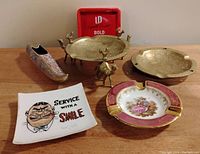 Shot showing six vintage ashtrays including ceramic, brass, and metal designs arranged on wood surface with the red LD Bold metal ashtray at the back.