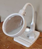Front view showing large magnifying lens and flexible neck with control button on the lamp arm, power adapter and cable coiled on base.