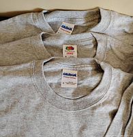 Close-up of neck tags showing two Gildan Heavy Cotton and one Fruit of the Loom Heavy Cotton labels on ash grey long sleeve t-shirts.