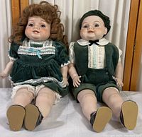 Both vintage matching porcelain dolls sitting side by side showing their overall attire and condition.