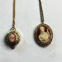 Photo of two gold tone cameo pendants with women's faces on chain necklaces, shown side by side on a white surface, displaying detailed relief and ornate metal surrounds.