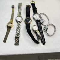 Photo of seven vintage wristwatches arranged in a row on a light fabric background. Watches include metal and leather bands, round and square faces in silver and gold tones.