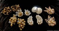 Six pairs of vintage earrings displayed on black fabric, showing a variety of styles including faux pearl clusters, gold-tone beads, and shell designs.