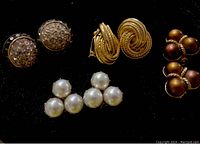 Photo of four pairs of vintage clip-on earrings arranged on black background showing different styles: rhinestone clusters, gold twisted ropes, brown pearl clusters, and large white faux pearls.