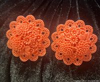 Close-up image of the pair of coral-colored celluloid floral clip-on earrings displayed on a dark textured fabric background, showcasing intricate flower and leaf details in rounded shape.