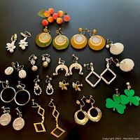 Overview of 15 pairs of vintage earrings with various materials, colors and clip-on/screw-back styles shown spread on black surface.