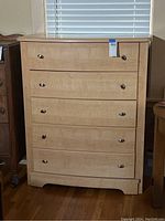 Front view of light wood dresser showing five evenly spaced drawers, each with two oval metal handles. The dresser has a smooth wood grain finish and stands on a short base with a slightly decorative cut-out bottom edge.