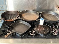 Overview of four cast iron skillets and one round cast iron trivet arranged on a stovetop. Skillets vary in size and shape; one with grill ridges and loop handles, others smooth and round or square shape. Surfaces show rust and wear.