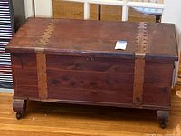 Front and side view of the cedar lined wooden blanket chest featuring decorative metal studded straps and a lock but no key, with four legs each on casters for mobility.